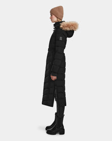 A person in profile wearing the Rudsak ELSIE M - L123517 Black/Natural women’s maxi down puffer with a fur-trimmed hood, black leggings, chunky boots, and a tan knit beanie, standing against a plain white background.