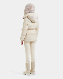 A person stands facing away, wearing the Rudsak EMBER M-M - L125524 Vanilla women's down puffer jacket with a fur-trimmed hood, paired with light neutral leggings and mid-calf boots, against a plain white background.