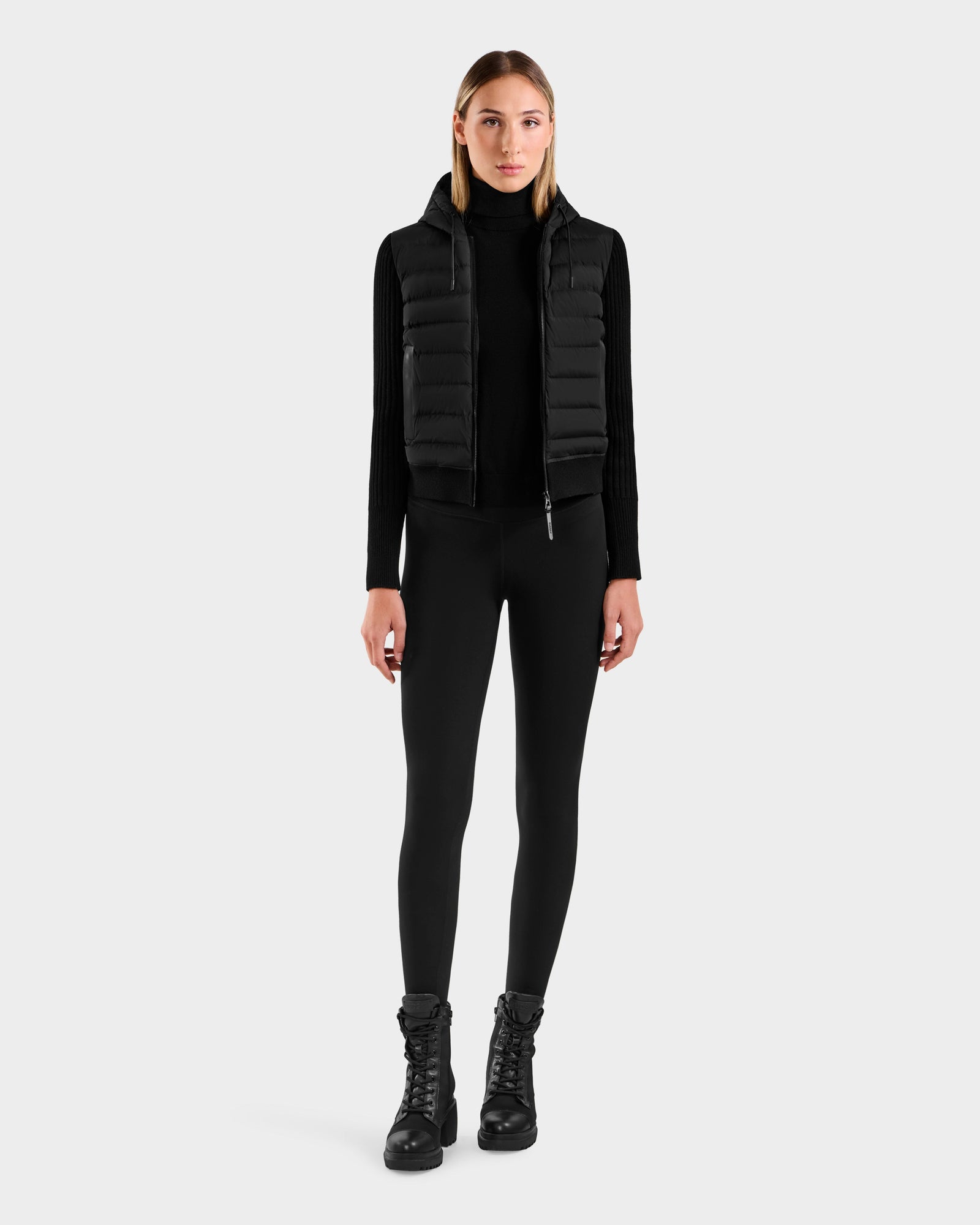 A woman faces forward wearing the Rudsak EMILY - L125515 Black hooded down bomber with a black turtleneck, leggings, and lace-up boots against a plain light gray background.