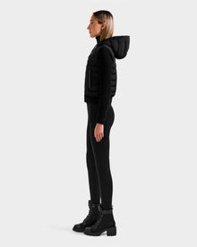 A woman stands in profile against a light gray background, wearing the Rudsak EMILY - L125515 Black women’s hooded down bomber with a black long-sleeve top, black leggings, and black lace-up boots.