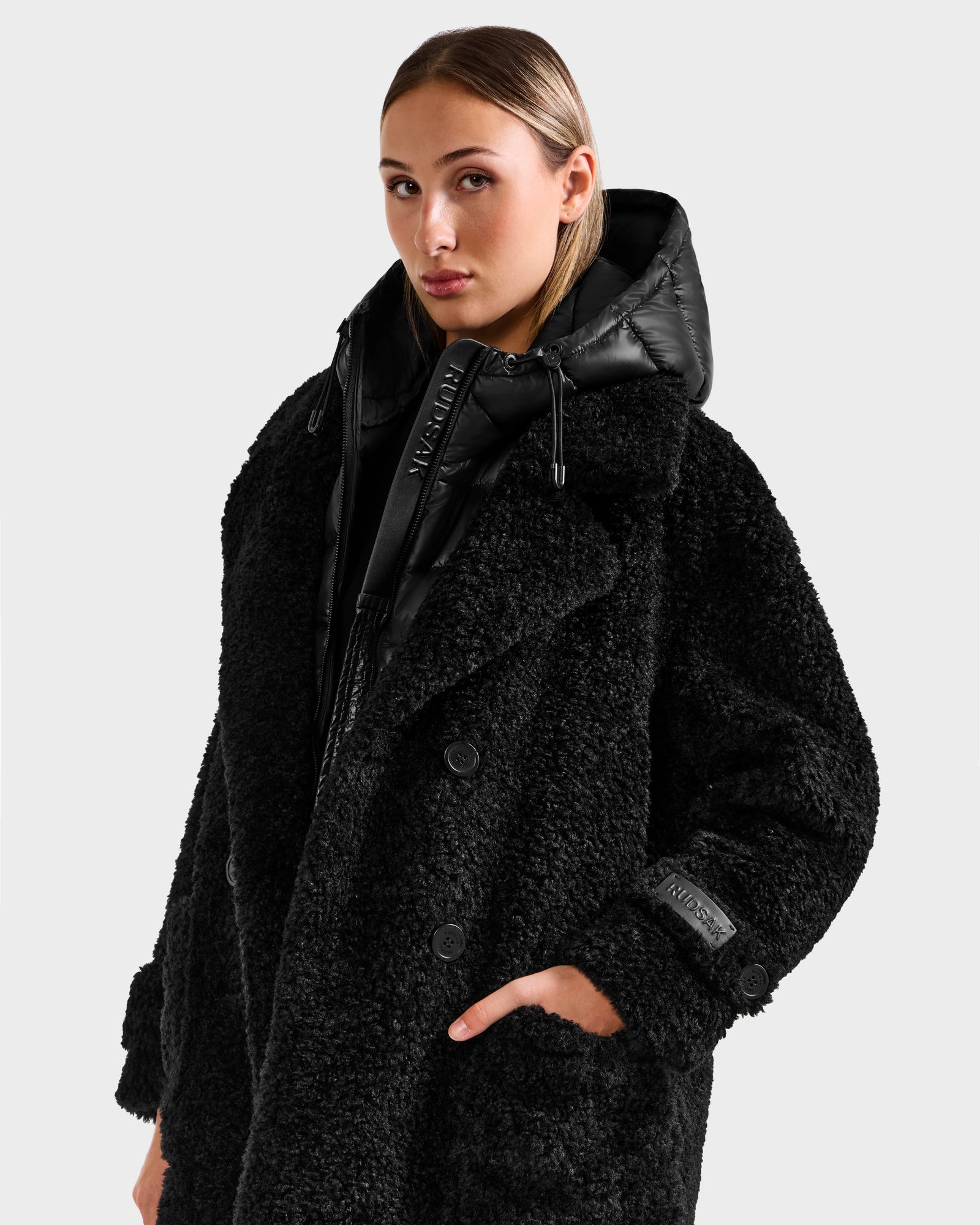A woman wears the Rudsak EMRYN - L125588 Black textured teddy jacket with a hood, one hand in her pocket, against a plain light background.