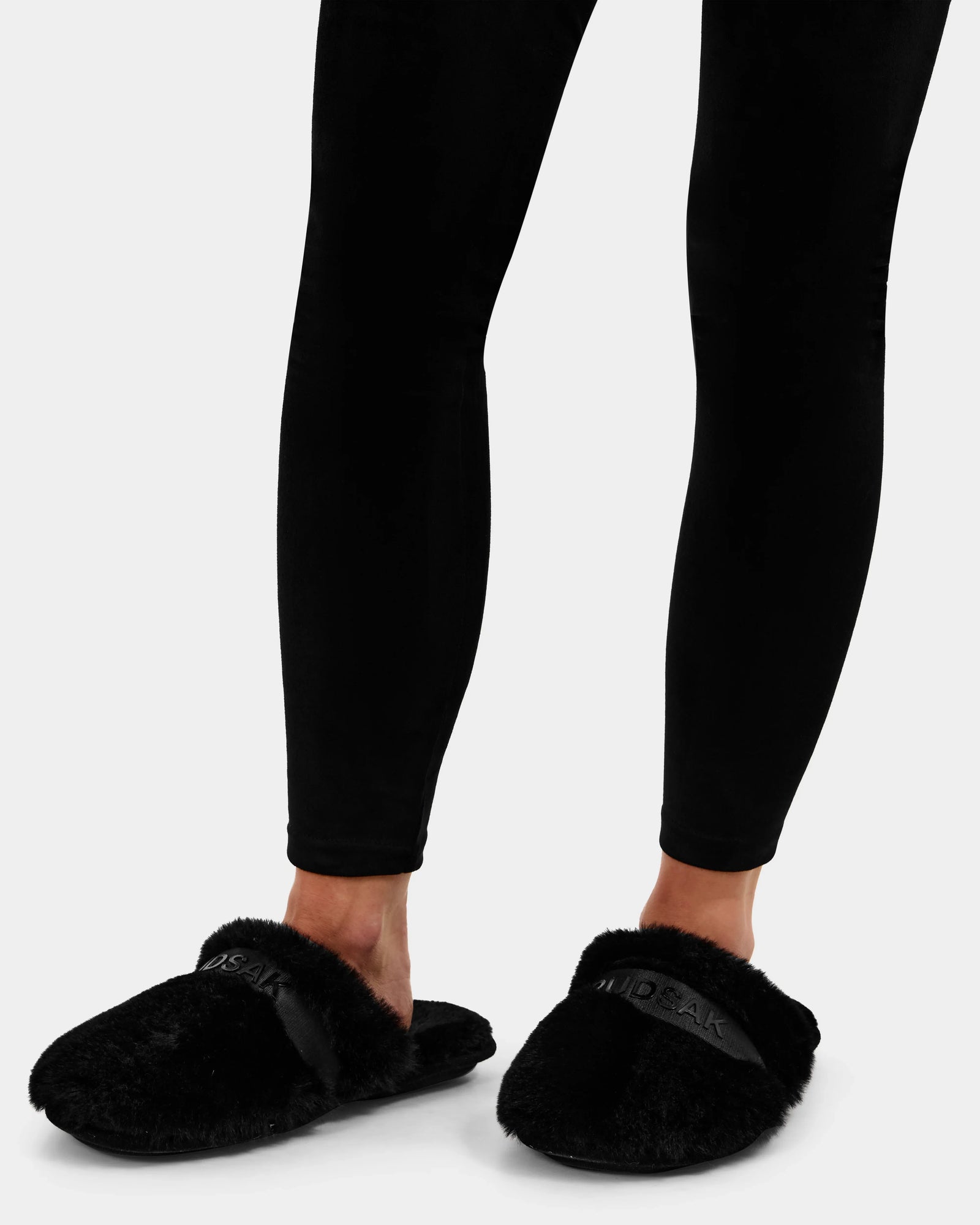 A person wearing Rudsak FLUFF - L223509 Black faux-fur slippers, with "NAKED" on the band, stands on a white background in black leggings—ideal cozy lounge slippers for relaxing at home.