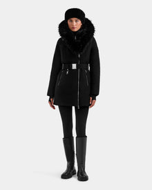 A woman faces forward in a Rudsak ISELLE - L125547 Black down parka with a large faux fur collar, silver zippers, and belted waist, paired with a black fur hat, leggings, and tall boots. The background is plain white.