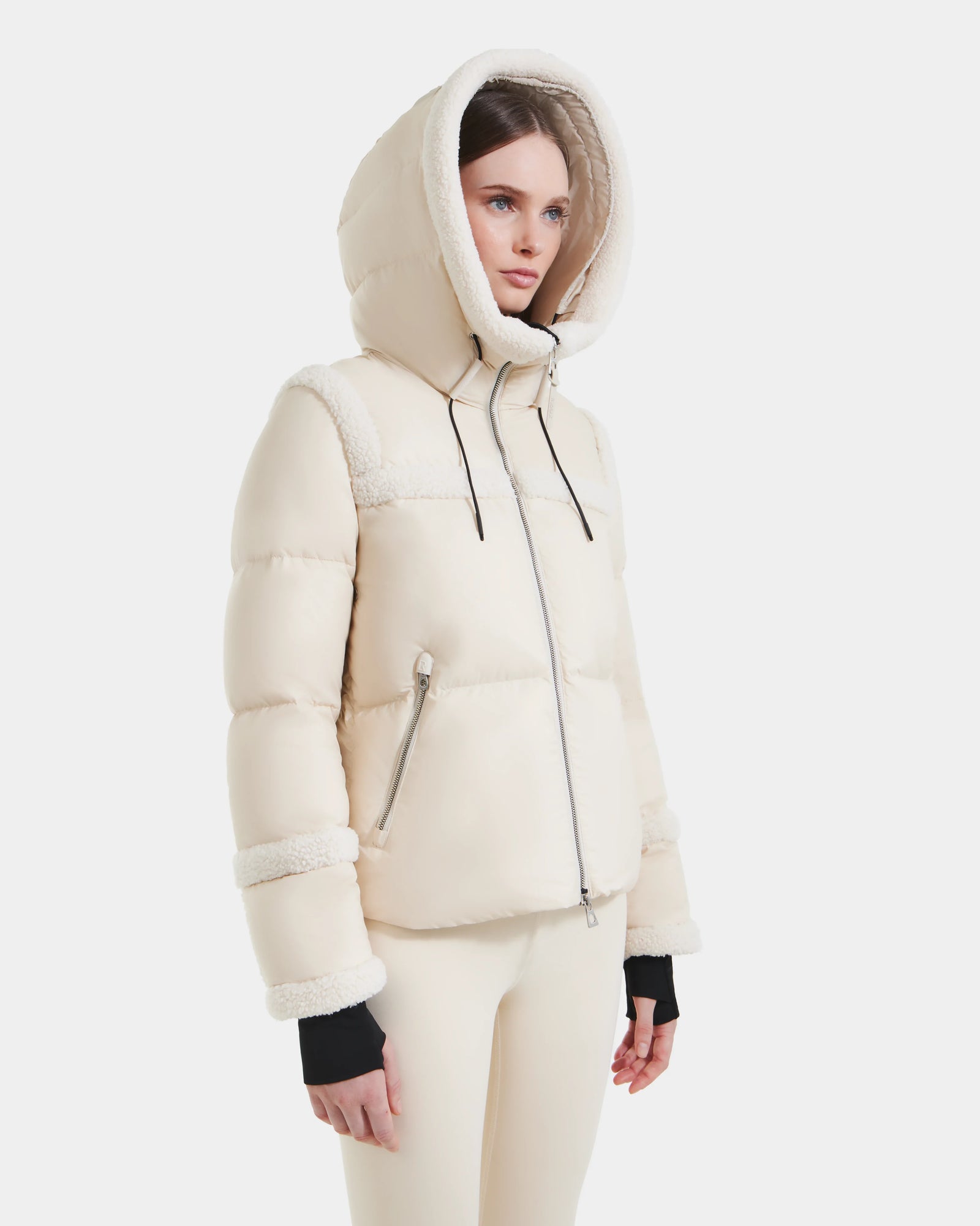 A woman wears the Rudsak JULIANA - L124509 Vanilla short down puffer jacket with a large hood and zippered pockets, paired with cream-colored pants against a plain white background.