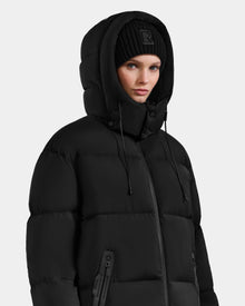 Wearing the Rudsak KARLEE XL - 8125683 Black women's maxi down puffer jacket with the hood up and a black knit beanie, she looks forward with a neutral expression against a plain light background—a winter essential.