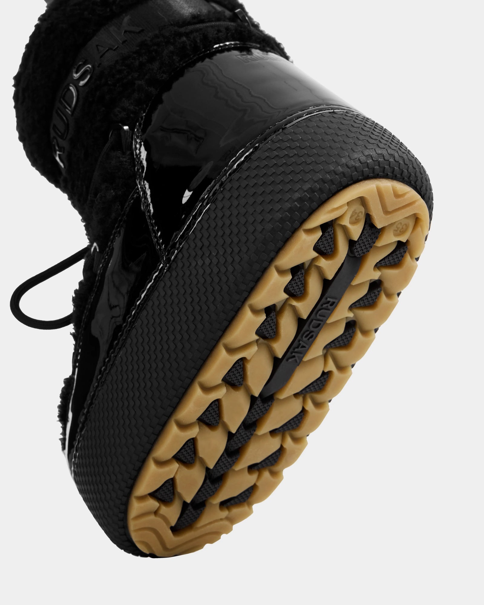 Close-up of the sole of a black, water-repellent Rudsak KATHY Y - 1224500 winter boot with tan, textured treads for grip; “RUDSAK” appears on the sole and side, featuring glossy and fuzzy materials inspired by classic moon boots.
