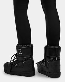 A person is shown from the back in black leggings and Rudsak's KIKA - L224506 Black women’s winter boots, featuring a shiny finish, chunky rubber sole, and laces, set against a plain light background—perfect for winter outings.