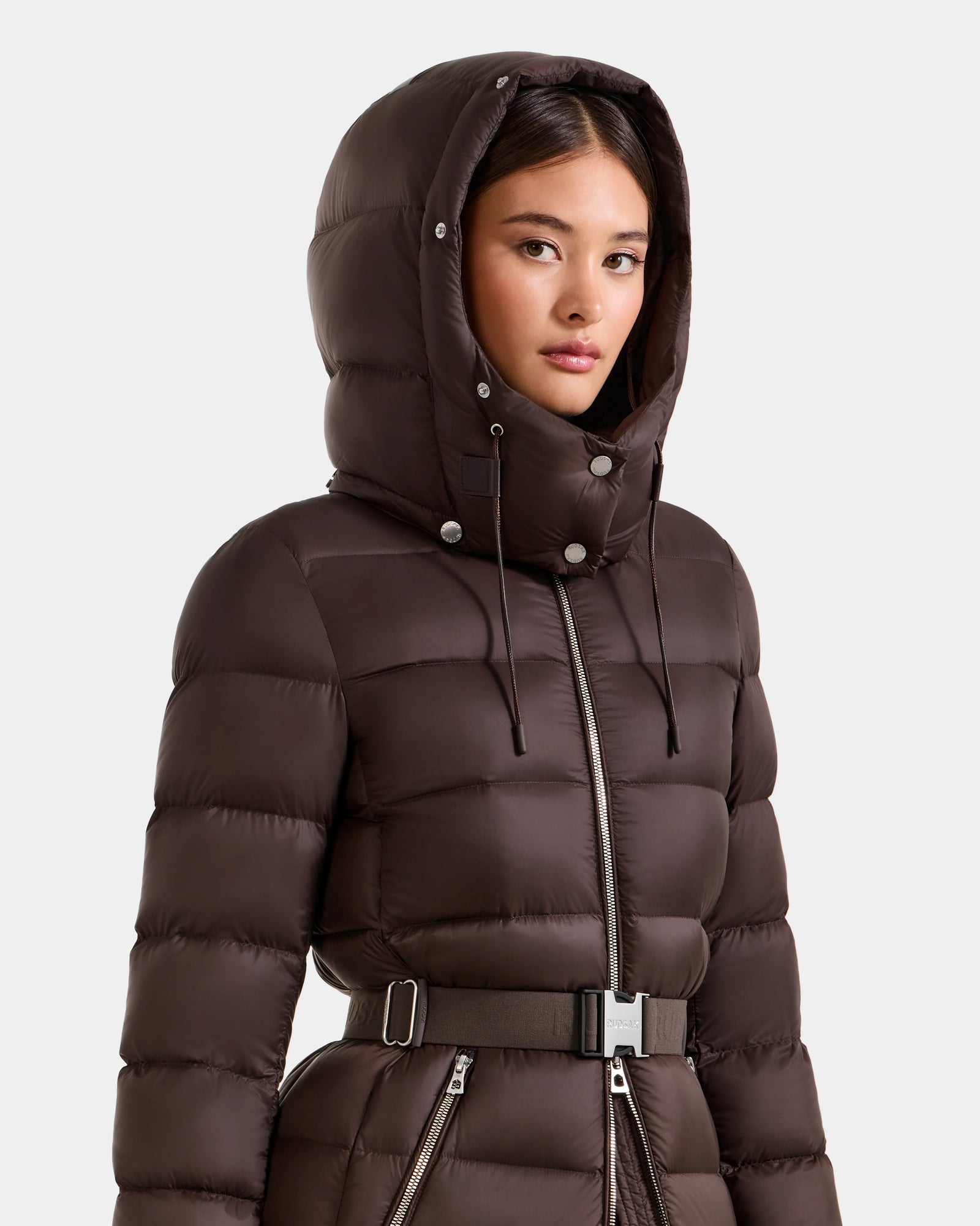 A woman models the Rudsak LILITH L - L125528 Espresso, a dark brown maxi down puffer coat with RDS-certified fill, hood, front zipper, and removable belt, standing against a plain light background.