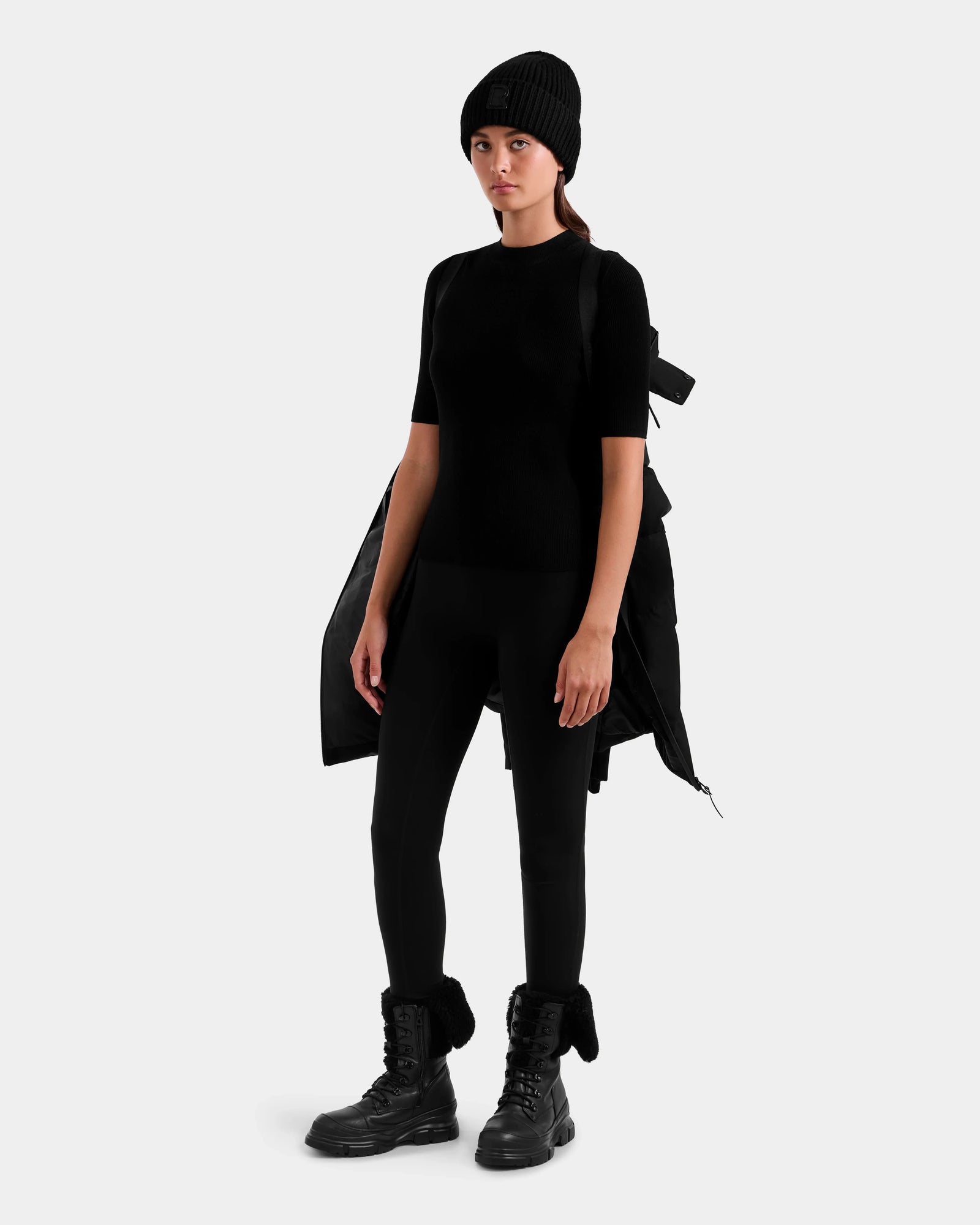 A woman stands against a plain light background, wearing a black beanie, short-sleeve top, leggings, and lace-up boots. She holds the Rudsak MIRIAM MW - L125604 Black women’s down puffer over her shoulder.