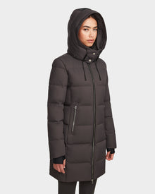 A woman in the Rudsak MIRIAM - L123544 Grey womens down puffer, featuring a weather-resistant shell, front zipper, zippered side pockets, and black hood drawstrings, stands slightly turned against a plain white background.