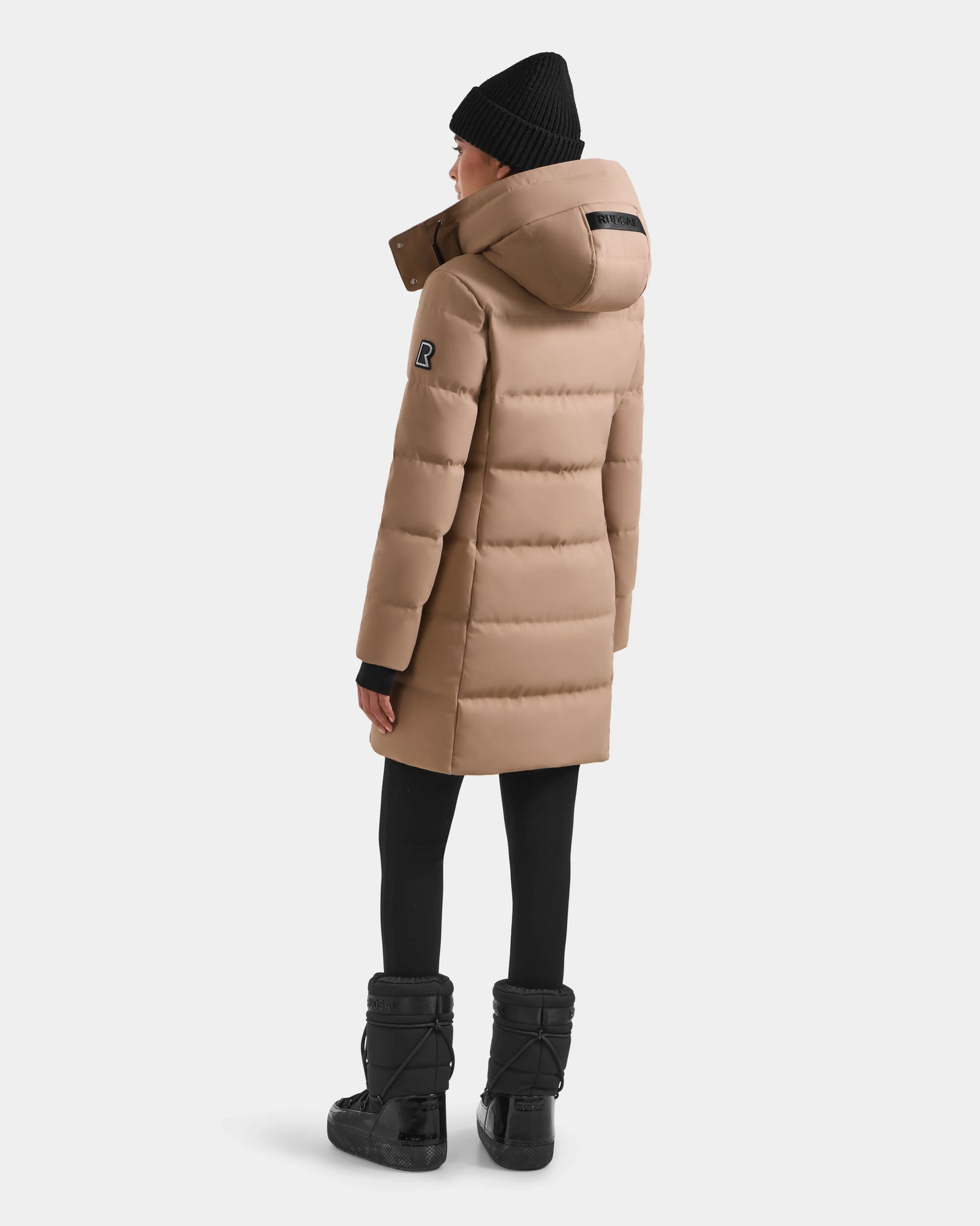 A person wearing the Rudsak MIRIAM - L123544 Sand women’s down puffer coat faces away, displaying the back of the weather-resistant design against a plain light background. She pairs it with black leggings, winter boots, and a black knit beanie.