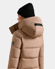 A person is shown from behind wearing the Rudsak MIRIAM - L123544 Sand weather-resistant puffer jacket with a large hood, black beanie, a black text patch on the hood, and a logo patch on the upper sleeve.