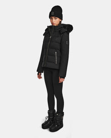 A woman wearing the Rudsak MIRRY M - L124533 Black quilted down coat with fur-trimmed hood, plus a black knit beanie, leggings, and winter boots stands against a plain white background.