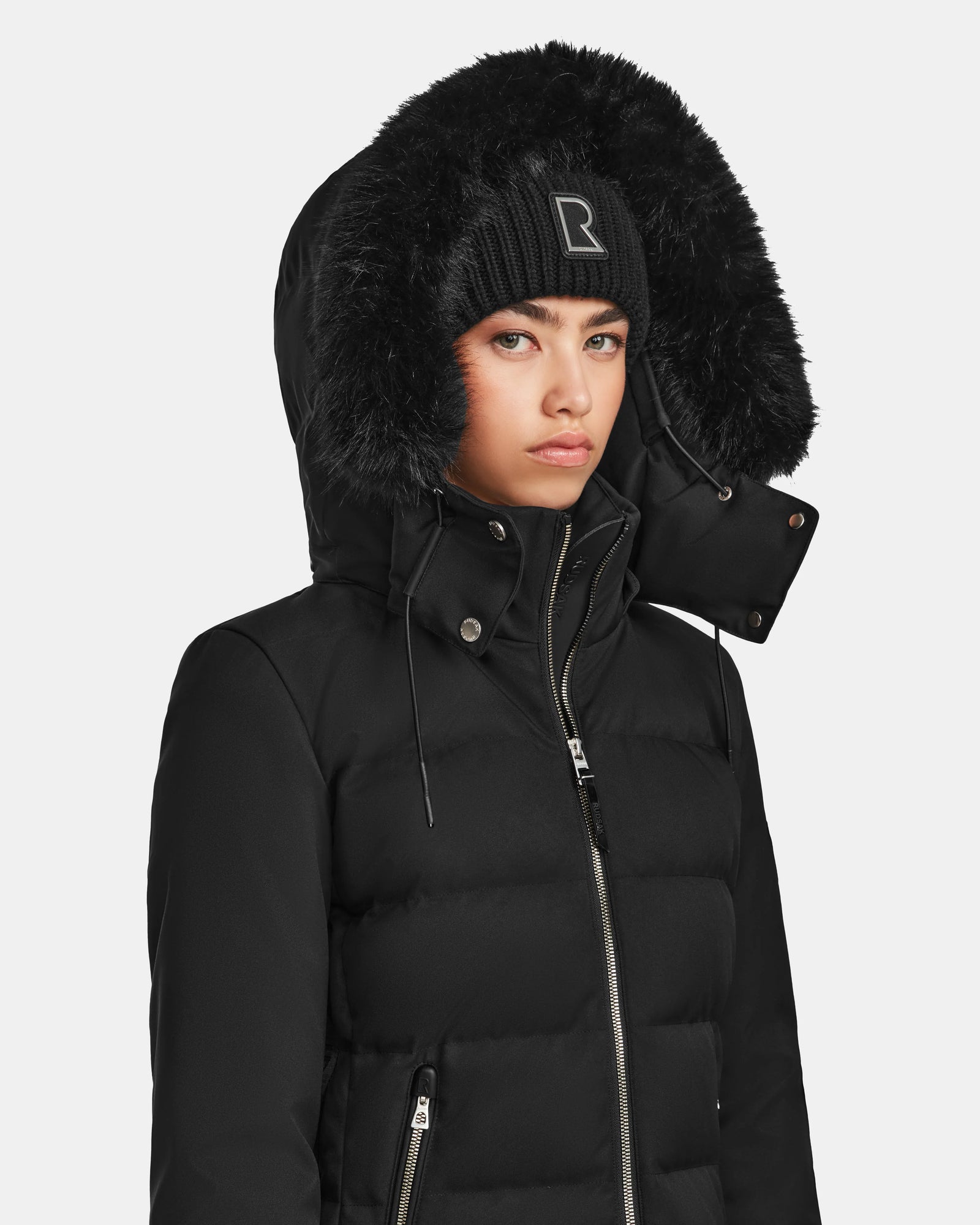 A person wearing a black Rudsak MIRRY M - L124533 winter coat with a fur-lined hood and a black knit beanie with a rectangular logo, looking straight ahead with a neutral expression.