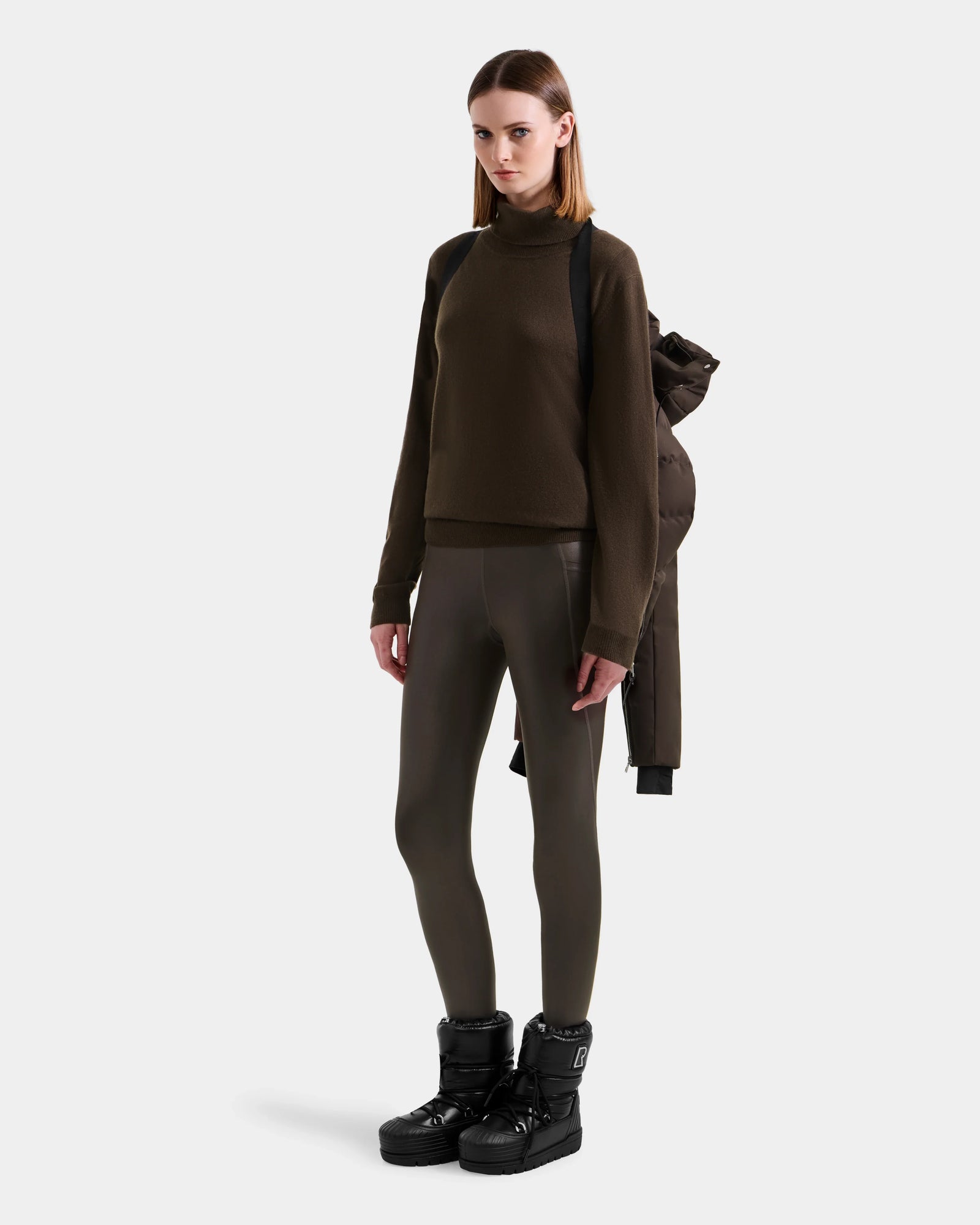 A woman faces forward against a white background, wearing an espresso brown turtleneck, shiny leggings, black boots, and the Rudsak MIRRY M - L124533 Espresso women's quilted down coat slung over her shoulder like a backpack.