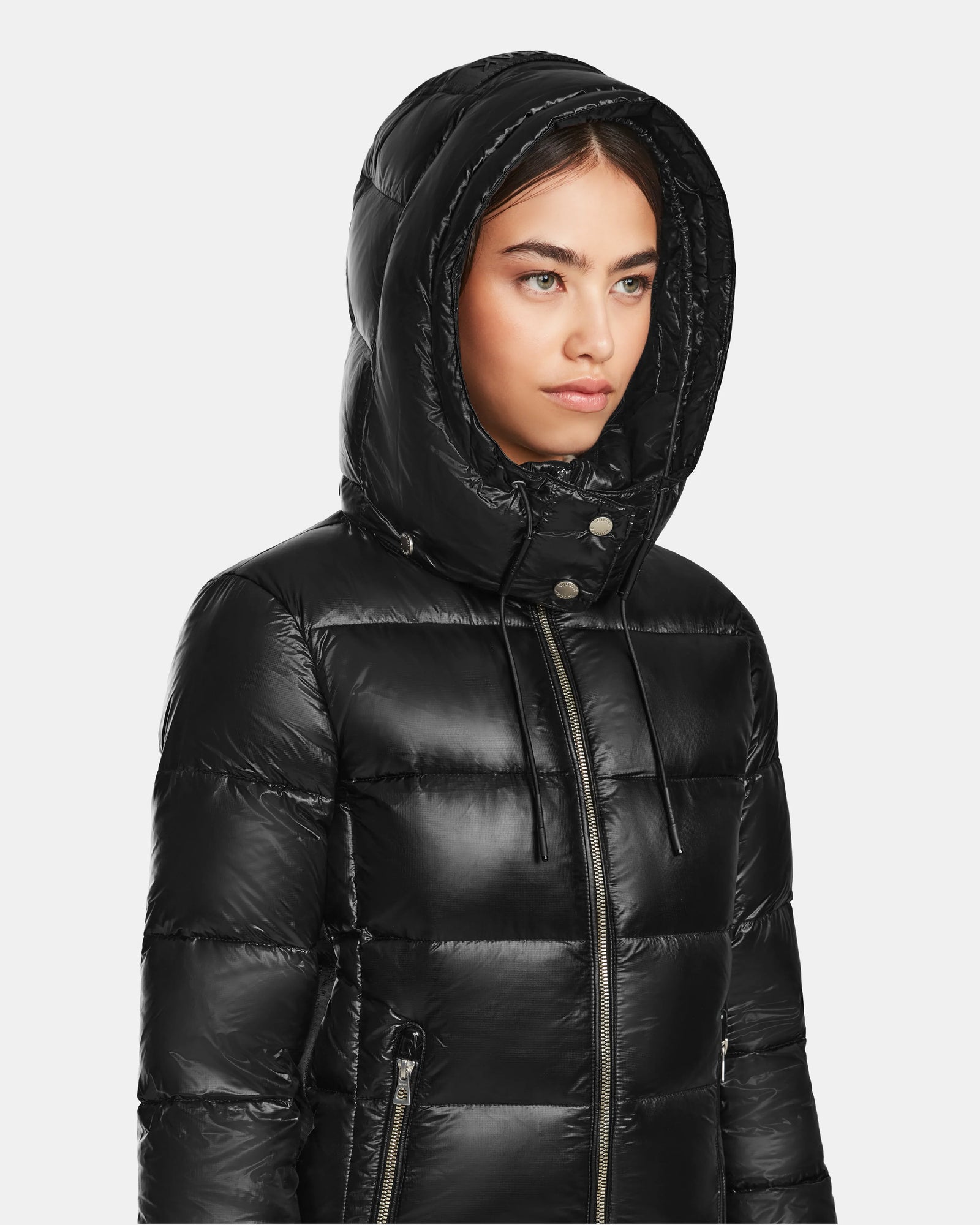 A woman in a shiny black Rudsak MIRRY S - L123545 quilted down coat with the hood up, glances to the side against a plain light background.