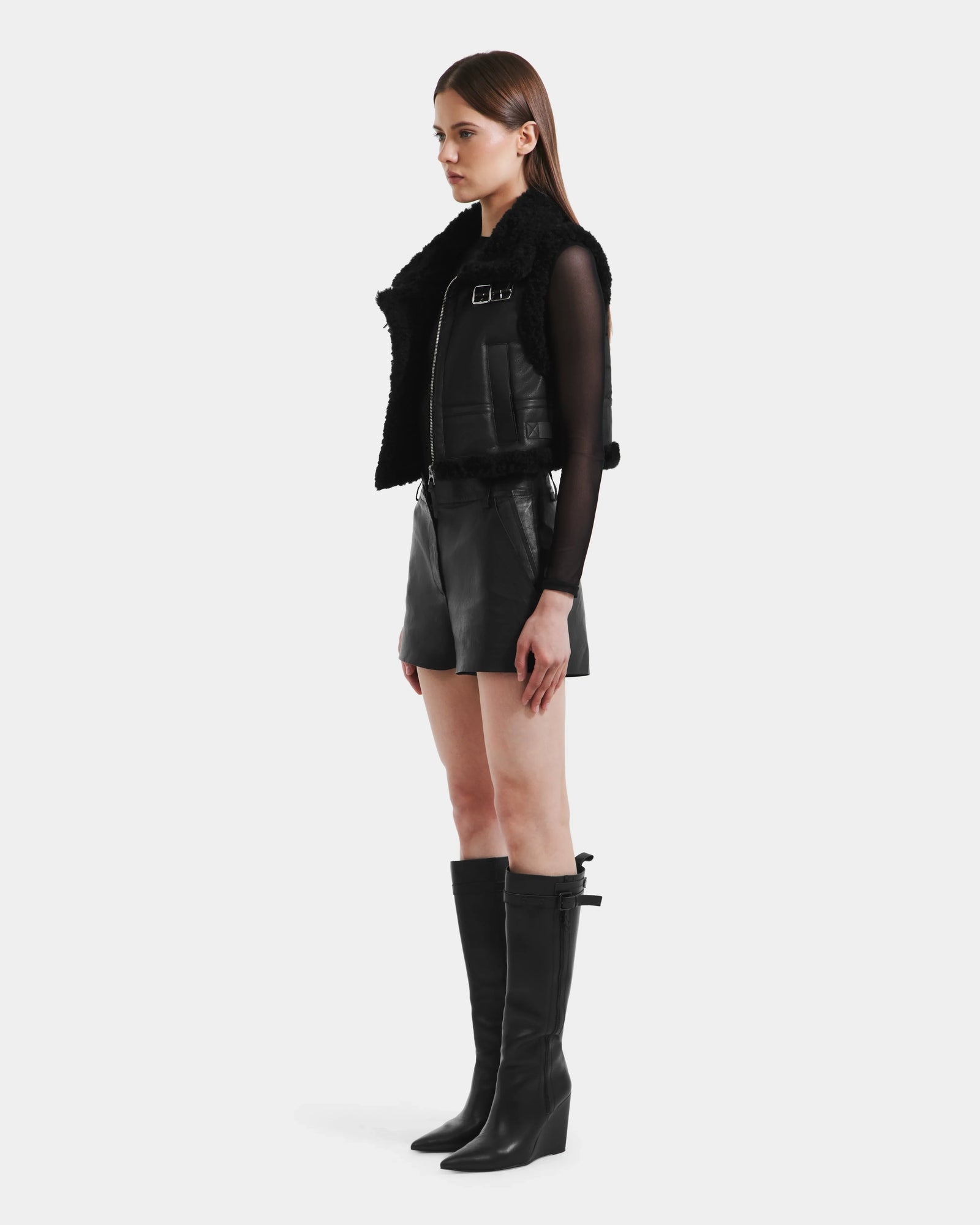 A woman in profile wears the Rudsak PAISLEY - L125516 Black faux shearling vest with a black leather mini skirt, black knee-high boots, and a sheer black long-sleeve top against a plain light gray background.