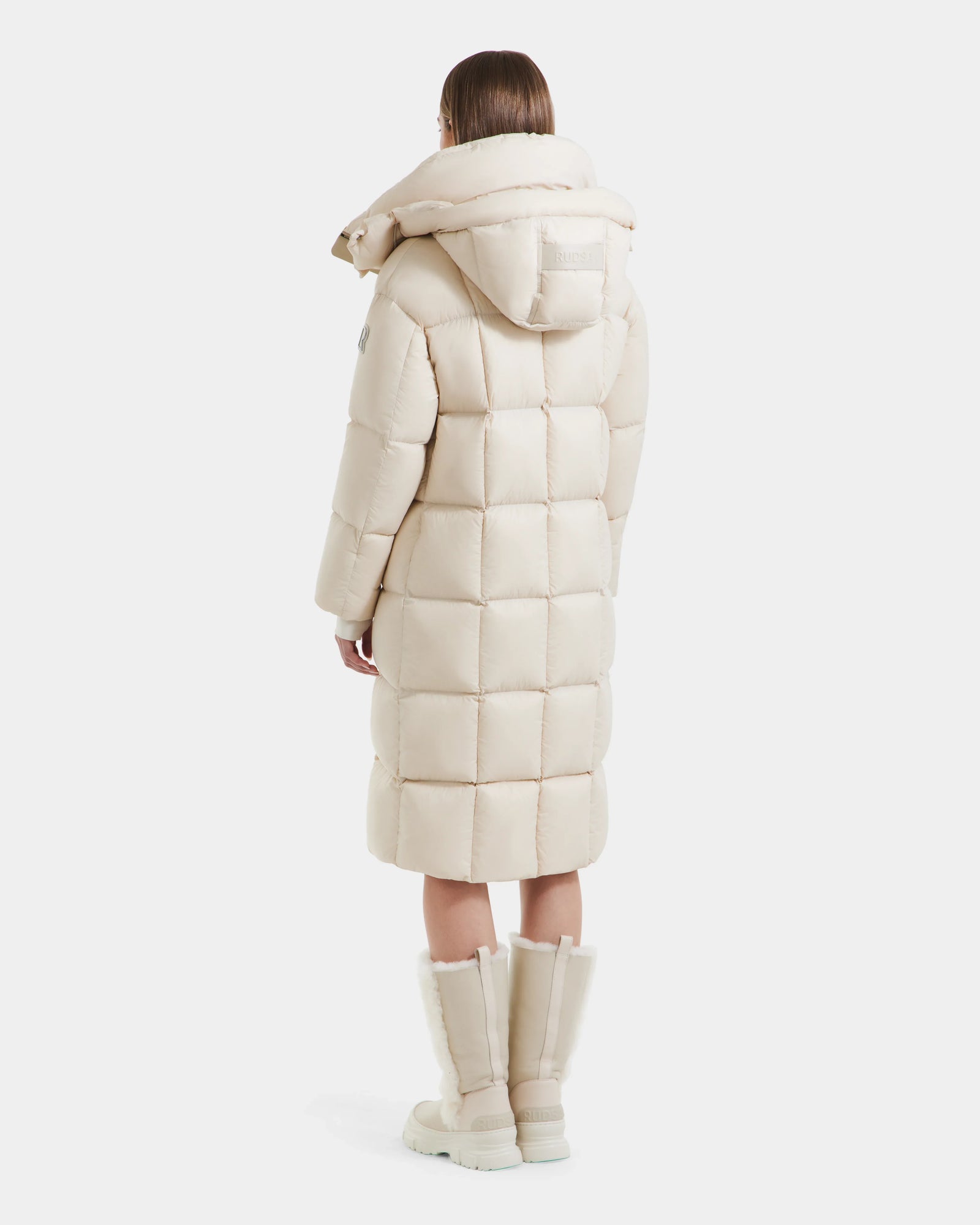 A person wearing the Rudsak SASHA - 8125686 Vanilla women’s long down puffer coat with a large removable hood and matching knee-high boots stands facing away against a plain white background.