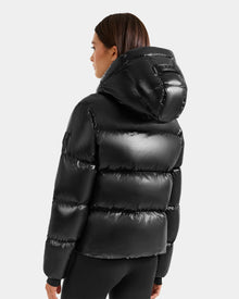 A woman with straight brown hair is seen from the back wearing the Rudsak STEFANIE S - L125585 Black women's short puffer jacket with hood, set against a plain light grey background.