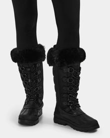 A person wearing black leggings and Rudsak WINDY - L224507 Black women's winter boots featuring faux fur trim, lace-up fronts, and sturdy rubber soles for cold-weather comfort.