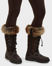 A person stands on a light background wearing dark brown leggings and Rudsak WINDY - L224507 Espresso women’s winter boots, featuring faux fur trim and lace-up fronts—ideal for cold-weather comfort.