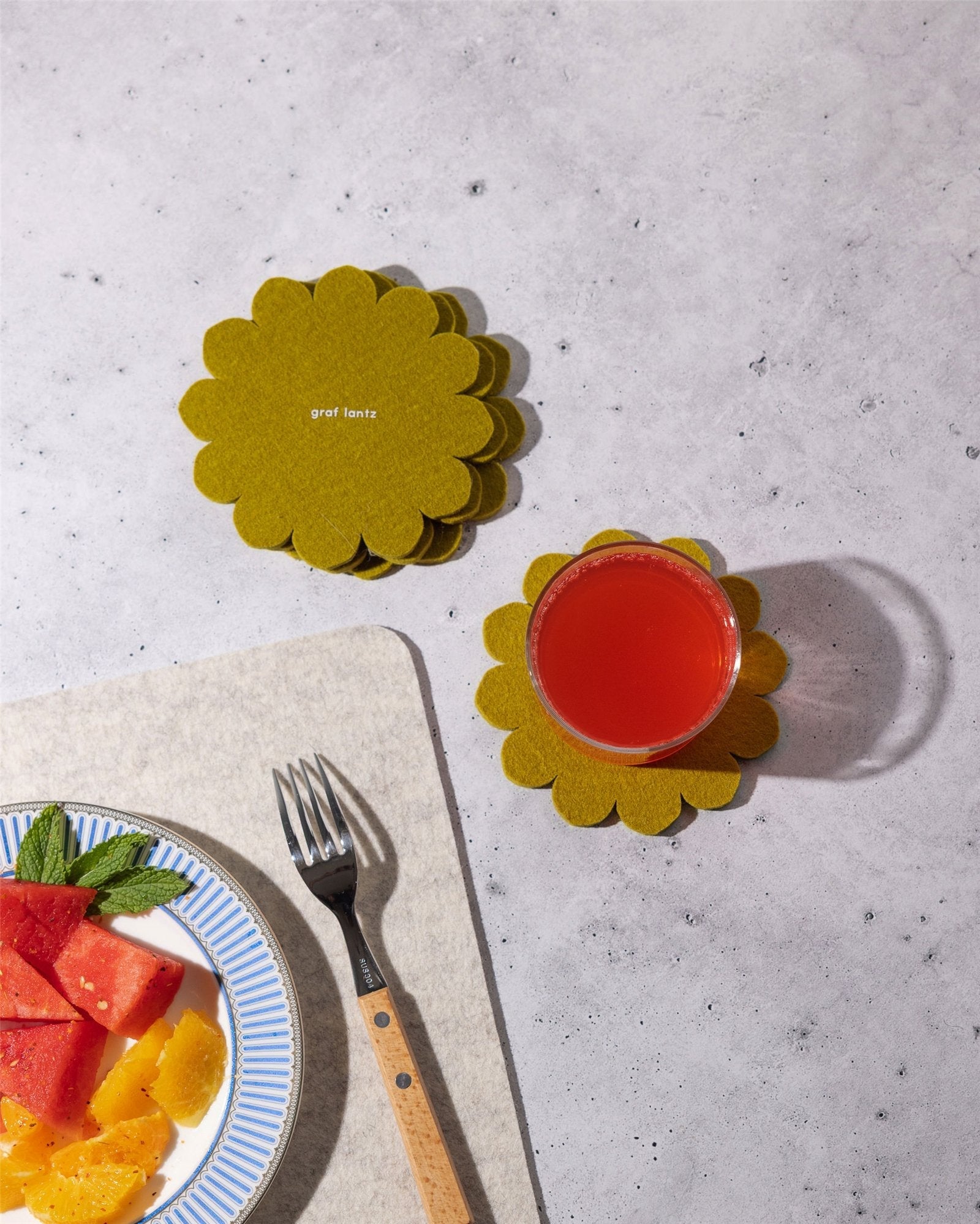 A glass of red juice sits on a yellow flower-shaped Merino wool coaster by Graf Lantz, inspired by Japanese floral crests. Nearby, more coasters are stacked for surface protection, with a plate of fruit and fork partially visible.