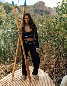 Woman holding two wooden sticks in a natural setting with mountains and greenery.