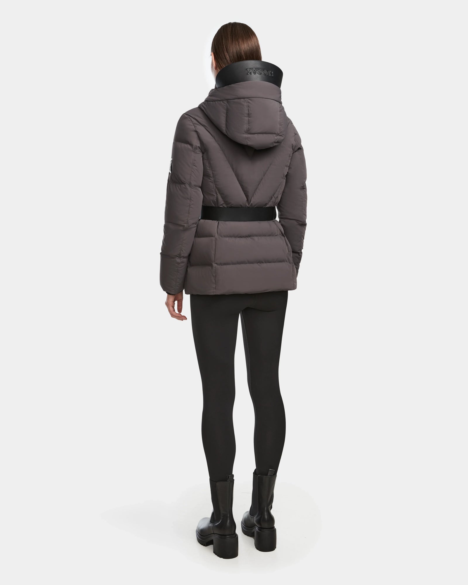 A person stands facing away, wearing the Rudsak ENZAH - 8124692 grey RDS-certified down jacket with a black belt, large hood, and high collar. The outfit includes black leggings and ankle boots, set against a plain white background.