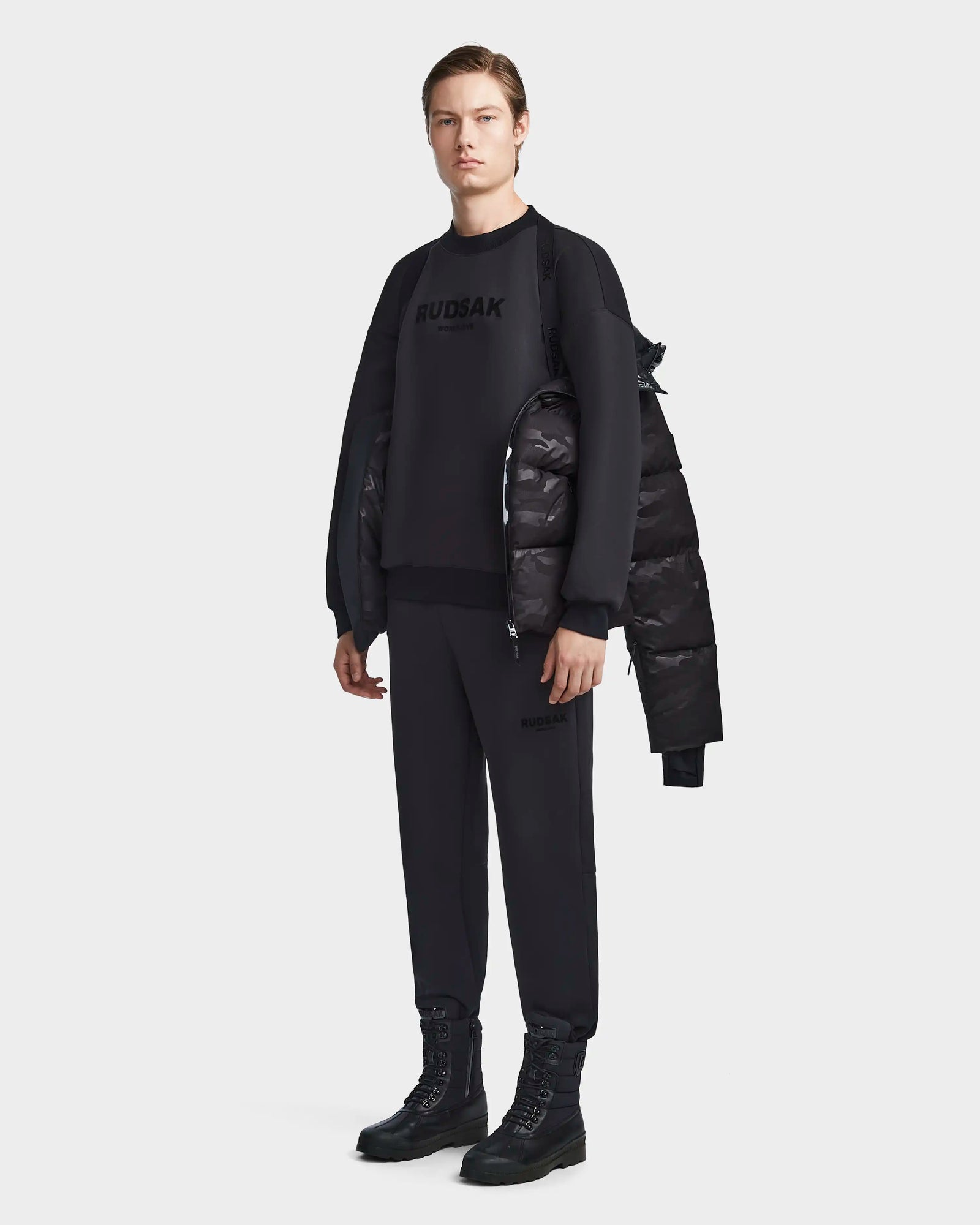A person faces forward against a white background, wearing the Rudsak RONNY M - M123512 Camo Black weather-resistant puffer jacket draped over one shoulder, with a black sweater, black pants, and black lace-up boots.