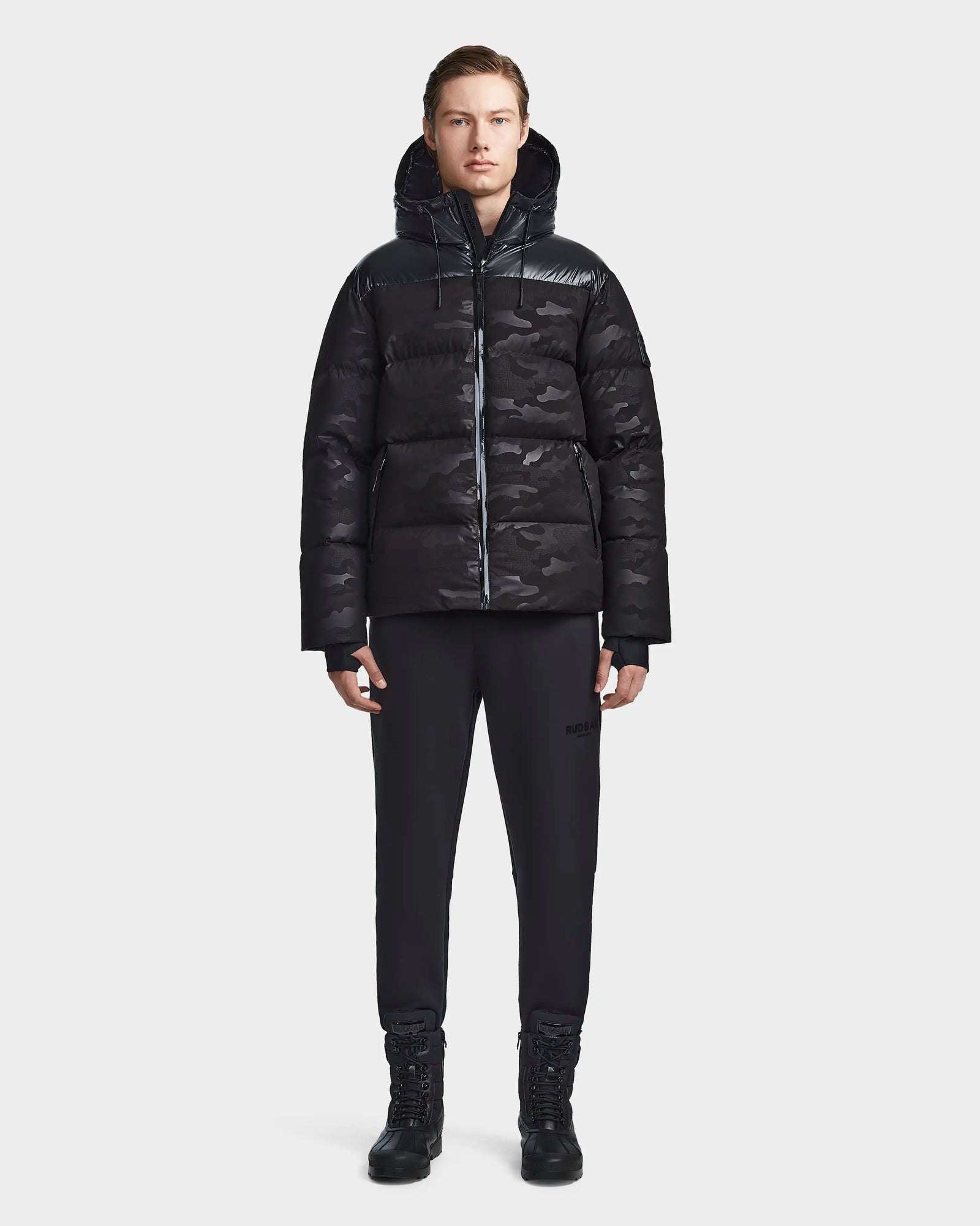A person faces forward wearing the Rudsak RONNY M - M123512 Camo Black weather-resistant hooded down puffer jacket with black pants and boots, set against a plain white background.