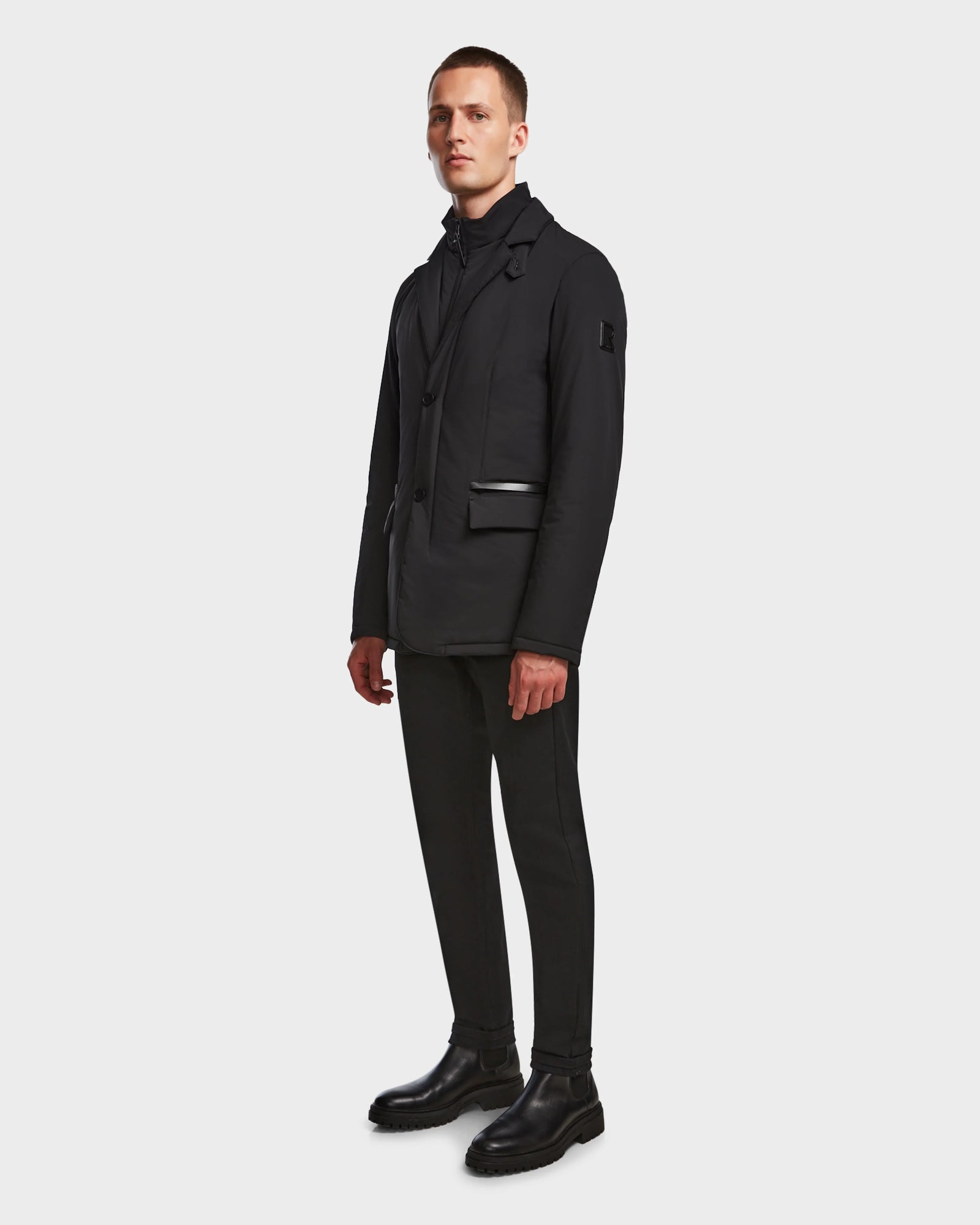 A man stands in front of a plain white background, wearing the Rudsak OTTO - 6124602 Black men’s blazer-style down jacket with eco polyfill, paired with black pants, a turtleneck, and boots.
