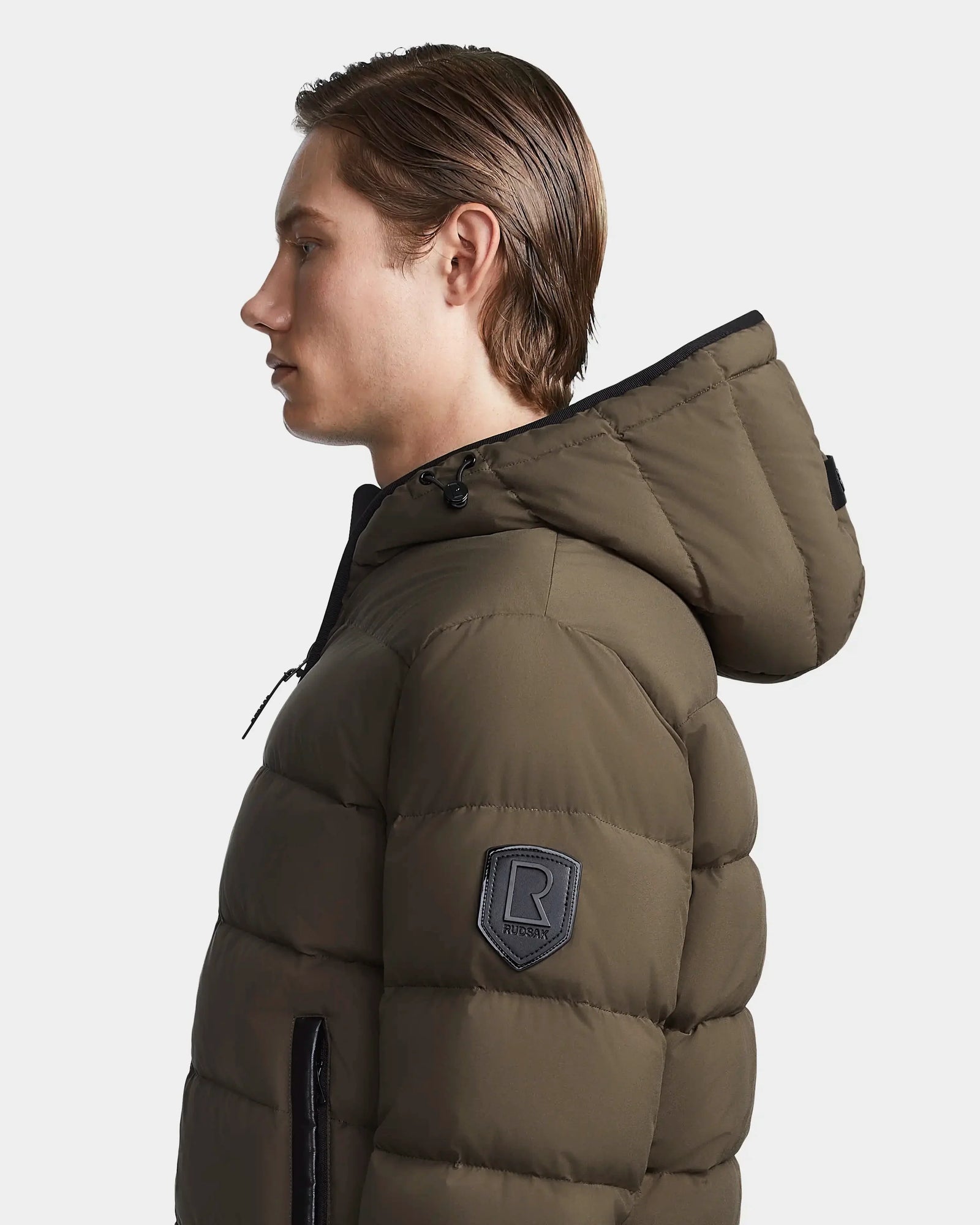 A person with straight brown hair is shown in profile wearing the Rudsak DEMIAN M - M123507 Light Olive men’s down puffer jacket, which features a black zipper and an upper sleeve patch with the letter R, set against a plain white background.