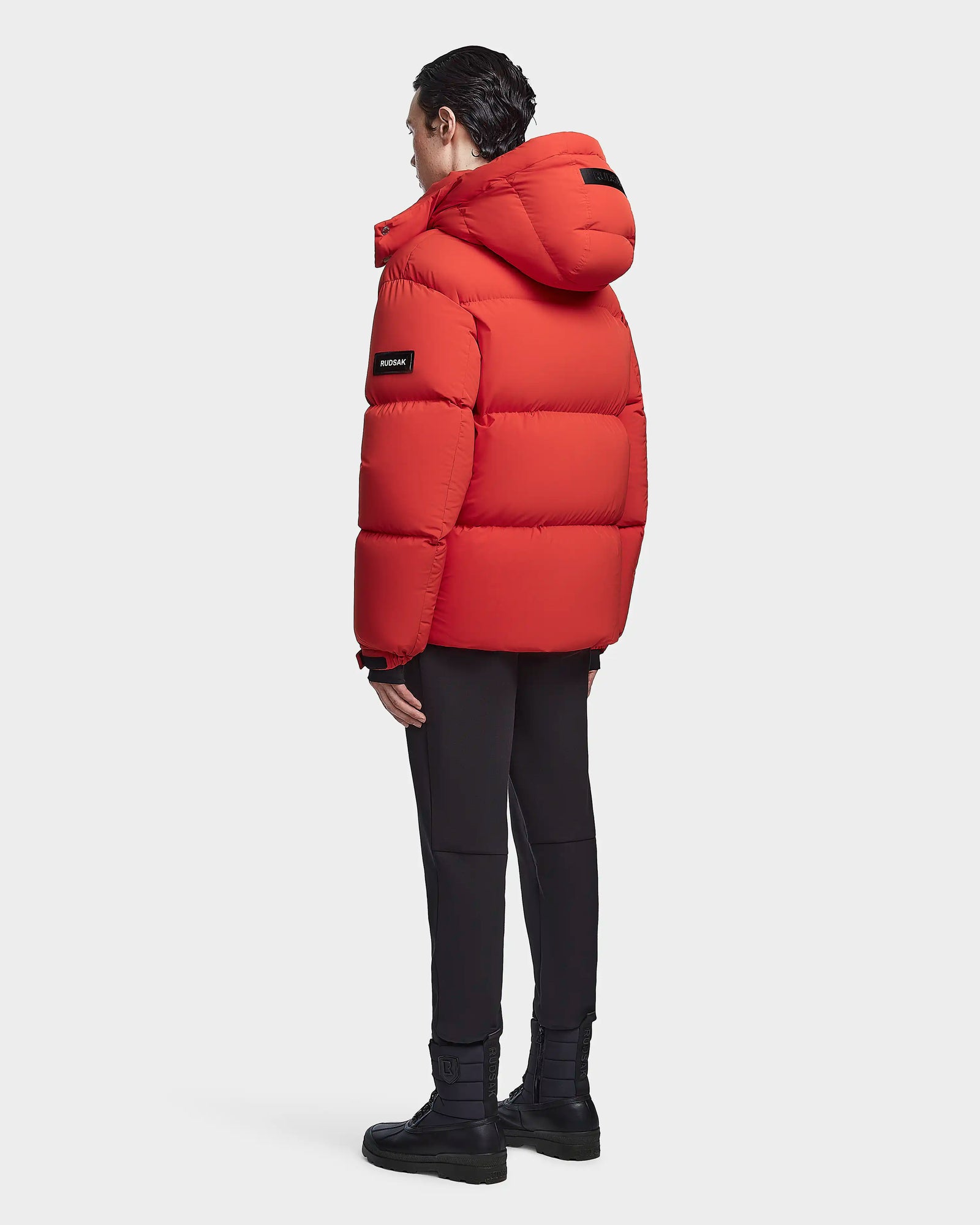 A person faces away, wearing the Rudsak VINCENT M - 6123510 Orange down puffer with hood and black patches, ultra-warm insulation, paired with black pants and boots against a white background.