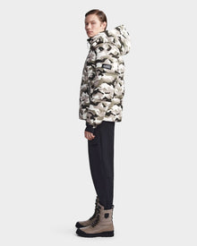 A person stands sideways against a plain light background wearing the Rudsak VINCENT M - 6123510 Camouflage men’s down puffer jacket in white, grey, and black, paired with black pants and tan lace-up boots.