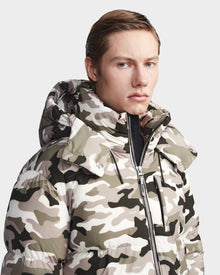 A light-skinned person with slicked-back hair wears the Rudsak VINCENT M - 6123510 Camouflage down puffer in beige and olive green, featuring a hood and water-repellent finish, posing with a neutral expression against a plain background.