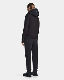 A person faces away wearing the Rudsak LAURENZO - M123514 Black neoprene jacket with removable hood, black pants, and chunky black boots, set against a plain white background.