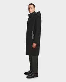 A man stands sideways against a plain white background, wearing the Rudsak DRAL - 6124503 Black mens wool jacket with a hood, paired with dark trousers and black boots.