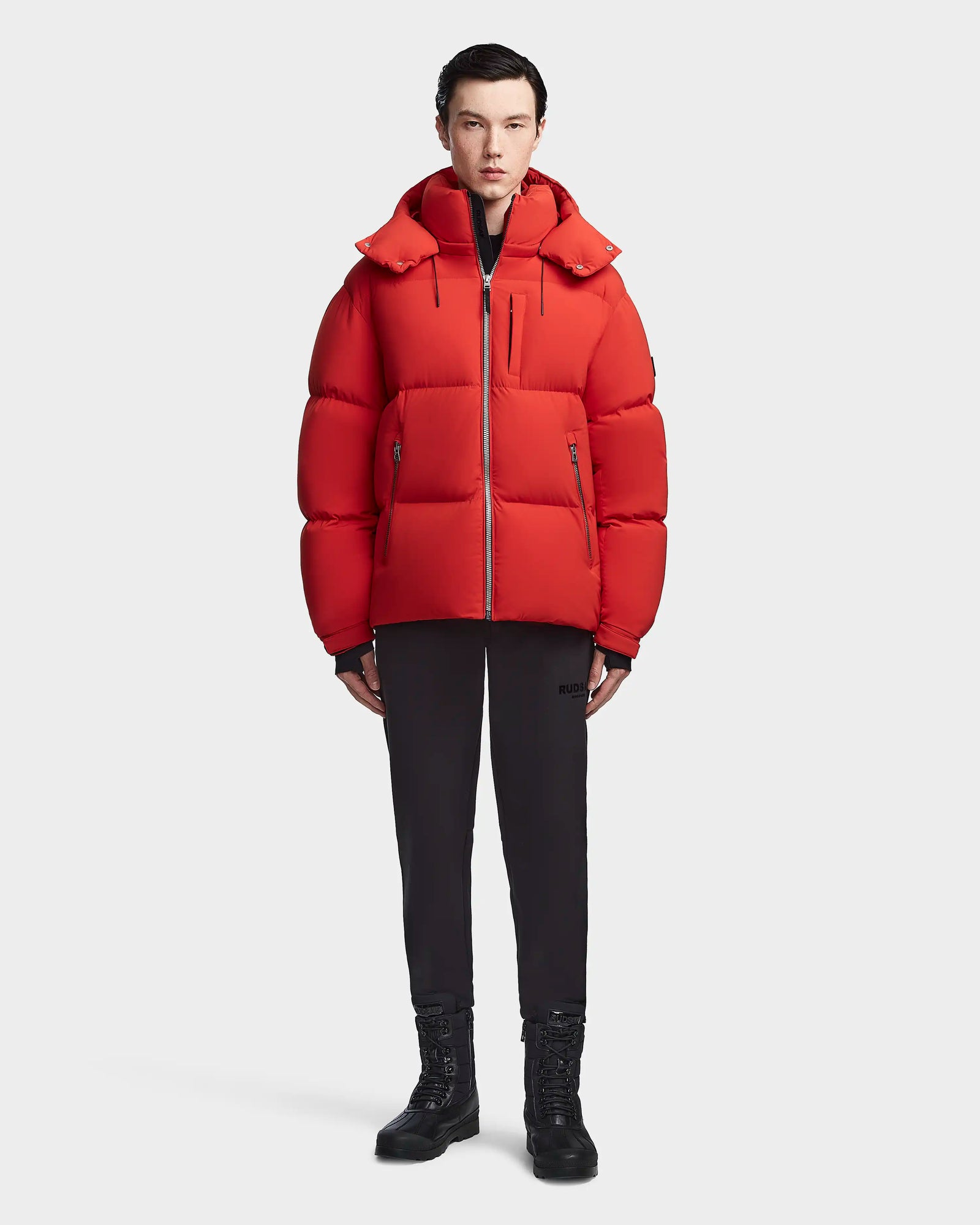 A person faces forward wearing the Rudsak VINCENT M - 6123510 Orange men’s down puffer, paired with black pants and black lace-up boots, set against a plain light background.