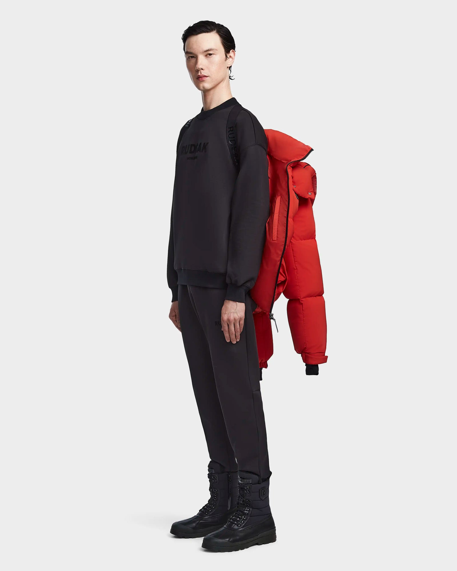 A person in black clothes and boots stands sideways with the Rudsak VINCENT M - 6123510 Orange men's down puffer, featuring ultra-warm insulation, draped over one shoulder against a plain white background.