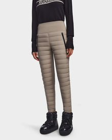 A person wearing Rudsak’s ARIL M - 8522514 Cement womens quilted pants with black zipper pockets, a black long-sleeve top, and shiny winter boots—apres-ski chic against a light backdrop.