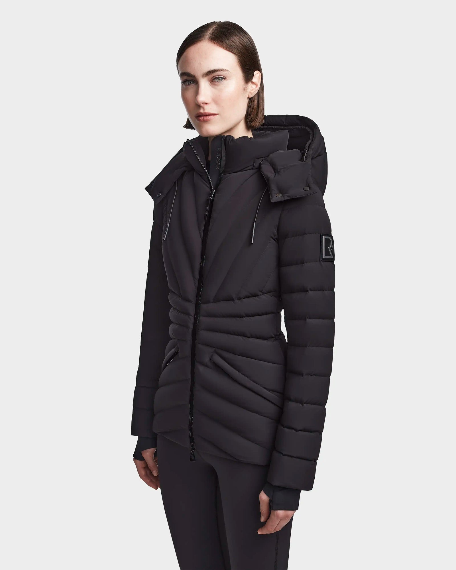 A woman with straight brown hair wears the Rudsak HANNA - 8123562 Black winter down jacket, featuring a fitted silhouette, hood, high collar, SMART STRETCH fabric, horizontal quilting, and subtle sleeve details against a plain light background.