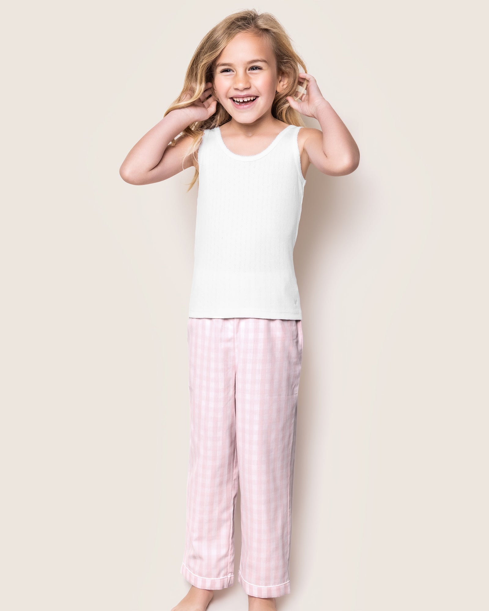 A smiling girl wearing Petite Plumes Kids Twill Pajama Pants in Pink Gingham and a white sleeveless top stands against a plain light background. Her long blonde hair cascades around her face as she gently rests her hands near her cheeks.
