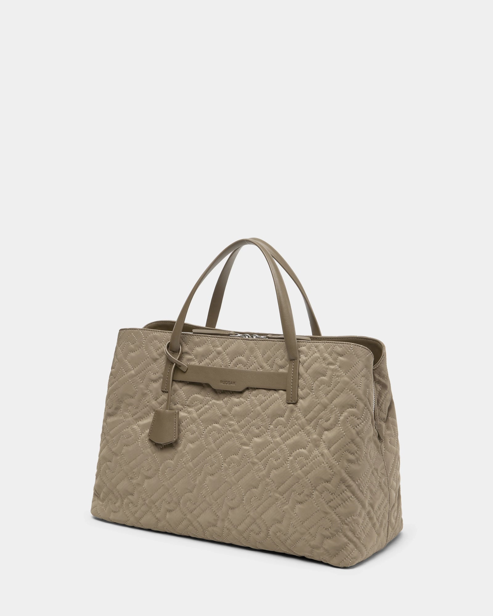 The Rudsak ASMINE - L325005 Almond is a beige quilted monogram tote with two top handles, an embossed pattern, and a small hanging tag, shown against a plain white background.