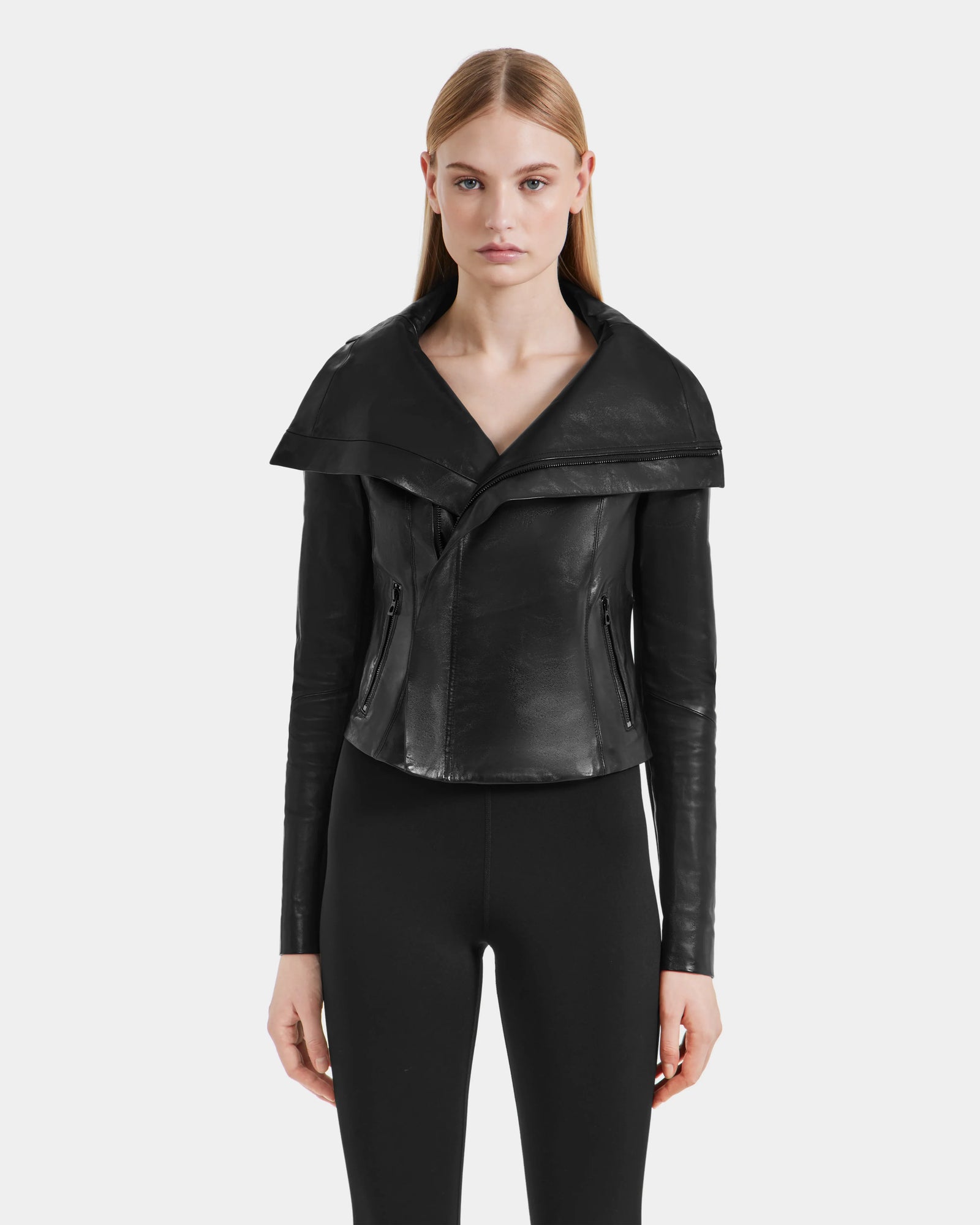 A woman with straight blonde hair wears the Rudsak AVRIL - 8124561 Black leather jacket, featuring a wide collar, paired with black fitted pants, standing against a plain light gray background.