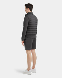 A man stands facing away against a plain light background, wearing Rudsaks RAGAB - 6124577 Grey mens hybrid jacket with lightweight down insulation, matching shorts, and white sneakers.