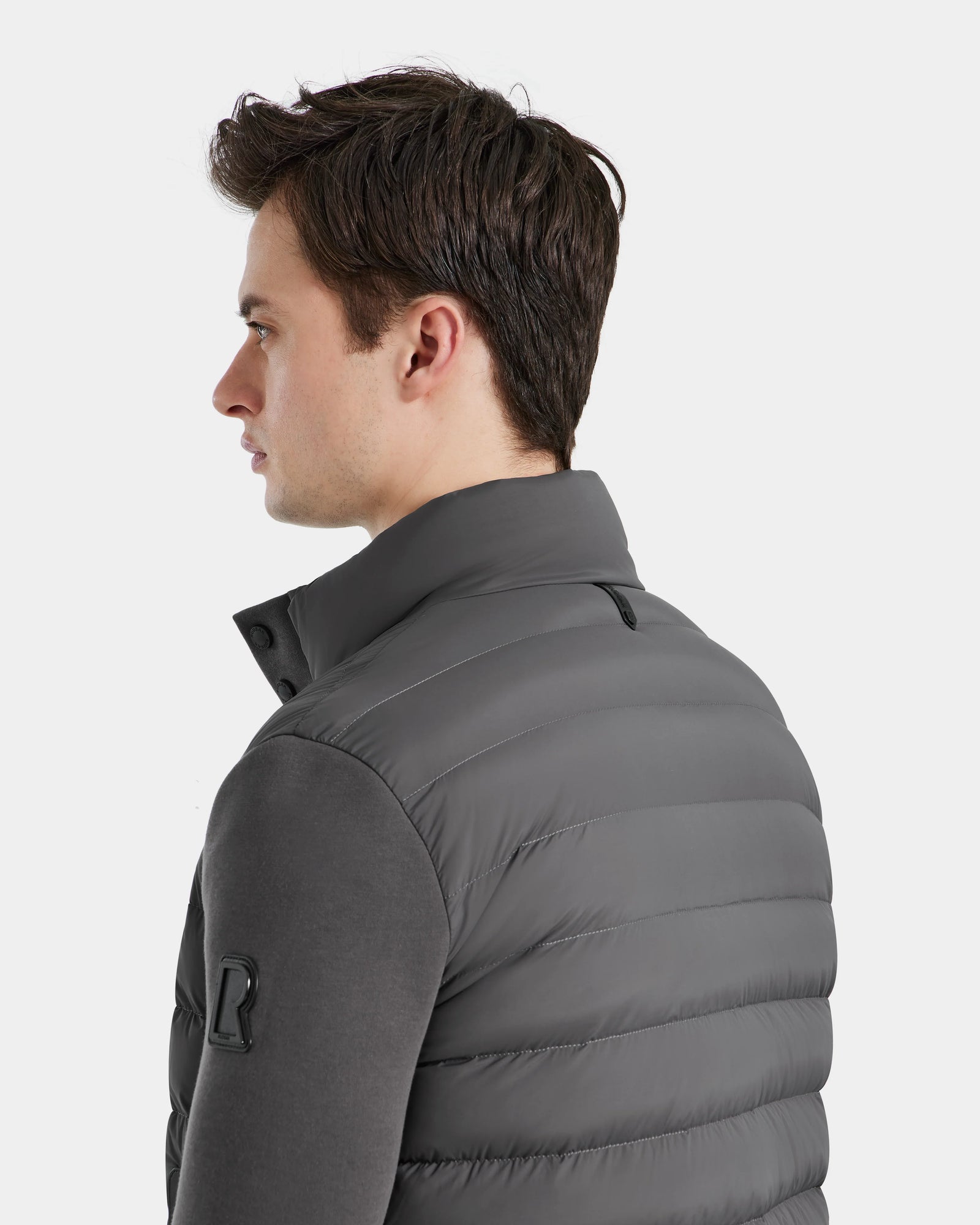 A man with short brown hair is seen in profile, wearing the Rudsak RAGAB - 6124577 Grey mens hybrid jacket with lightweight down insulation and a high collar over a dark long-sleeve shirt against a plain white background.