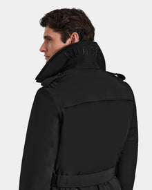 A man with short brown hair wears the Rudsak TIMOTHY - 6124589 Black mens trench coat. Its belted design, high collar with bold embroidered text, and water-repellent finish stand out against a plain light gray background.