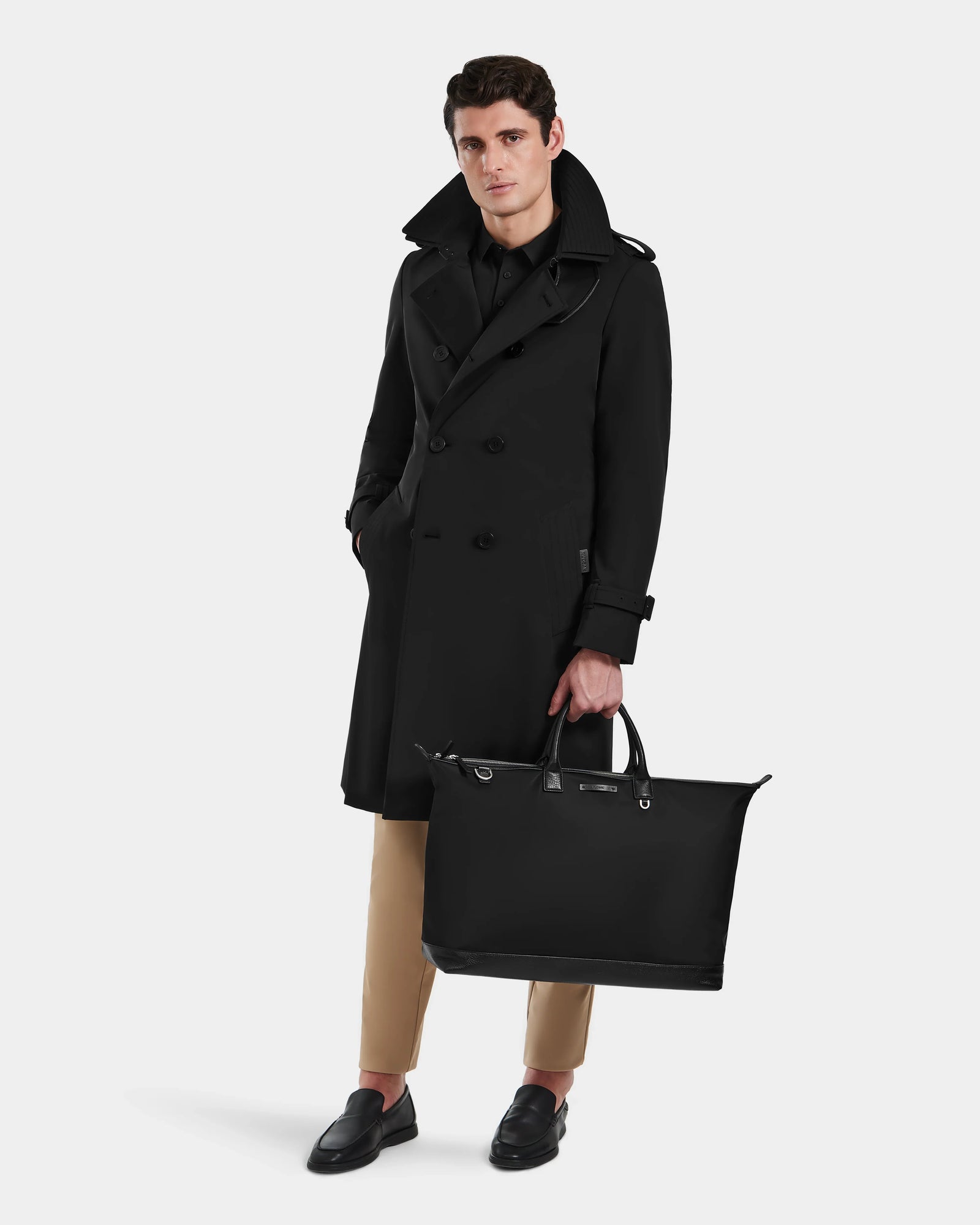A man wearing the Rudsak TIMOTHY - 6124589 Black knee-length trench coat and holding a bag.