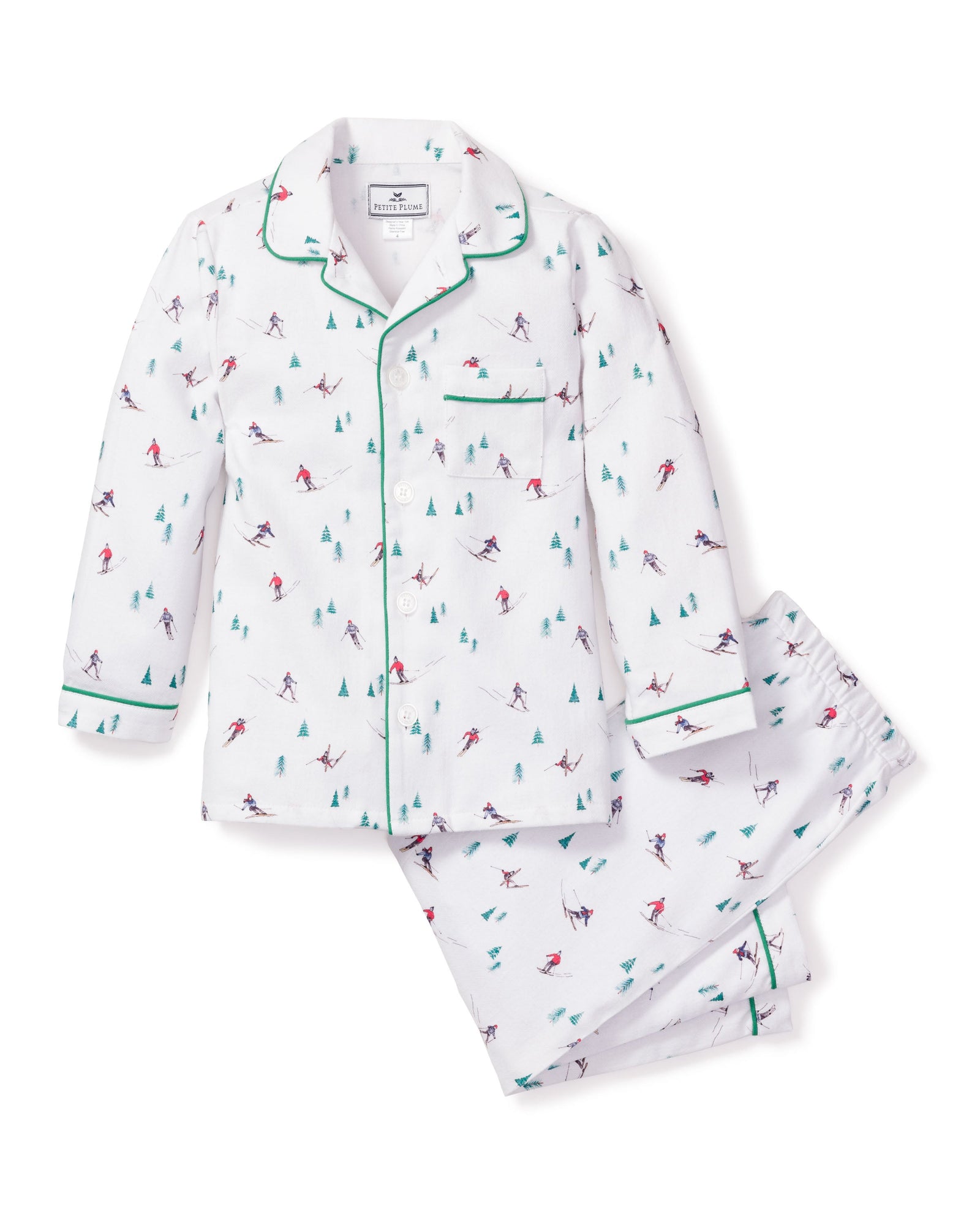 Petite Plumes Kids Flannel Pajama Set in Apres Ski features a white skiing pattern with small figures and trees. The button-down top has green piping on the collar and cuffs, while the matching pants have an elastic waistband. Made from flame-retardant fabric for added safety and comfort.