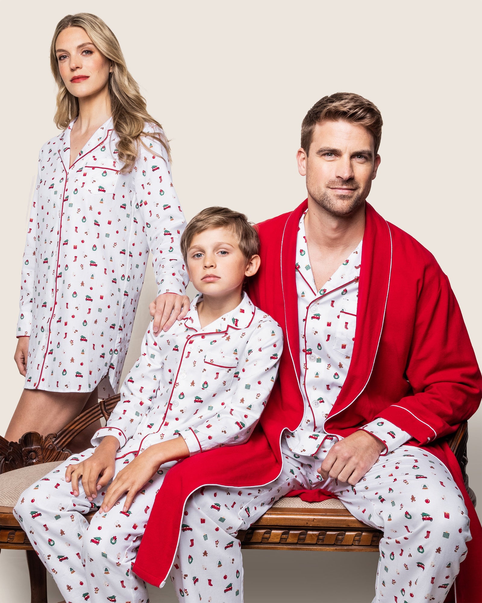 A family of three sits on a bench in matching Petite Plume Kids Twill Pajama Sets in Winter Nostalgia, adorned with festive prints and red trim. The man adds a red robe. A neutral backdrop enhances the cozy scene, poised for the gentle chime of Christmas bells to complete their cheerful image.